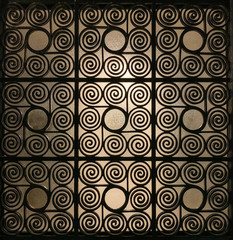 Wrought Greek iron gate grille, ornamented with elaborately twisted spirals and with frosted glass in the background, that is slightly illuminated. Metal sheet and barrier. Antique reproduction. Photo