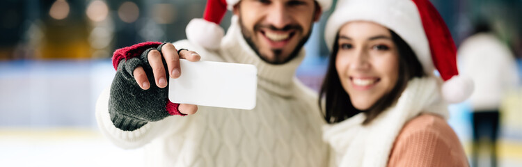panoramic shot of smiling couple in santa hats taking selfie on smartphone on skating rink at christmastime