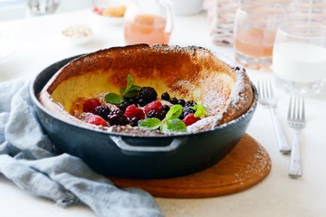 Fresh homemade dutch baby pancake with powdered sugar, fresh berries and mint for breakfast with a glass of milk and apple juice on a light background. Delicious sweet breakfast in a sunny interior. 