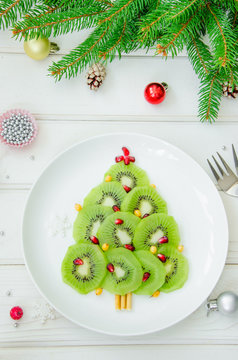 Kiwi Christmas tree on a white plate with pomegranate and sea buckthorn on a white wooden background. Christmas food idea. Healthy dessert.