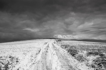 Dirt road leading to the top of the hill at wintertime, dramatic storm clouds in black and white.