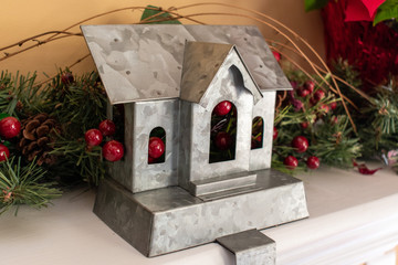 A metal vintage church surrounded by berries and garland sits on the mantle above the fireplace as decoration during the Christmas season. Photo is taken in a horizontal, landscape orientation