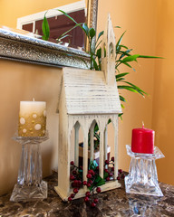 A wooden church with gold and red candles by its sides sits on an  entrance table as decoration for the Christmas season. Photo is taken in a vertical, portrait orientation.