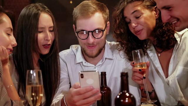Portrait shot of group of happy hipster students watching video, photo on the smart phone together while sitting at the wooden table in the modern bar, pub.