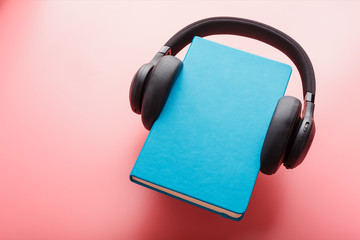 Headphones are worn on a book in a blue hardcover on a pink background, top view.