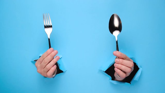 Female hands holding spoon and fork through torn blue paper wall. Gesture of eating dinning. Cooking, healthy homemade food concept. Copy space