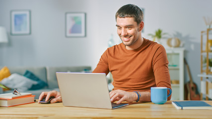 Portrait of Handsome Smiling Man Working on Laptop, Sitting at His Wooden Desk at Home. Man Browsing Through Internet, Working on Notebook from His Living Room.