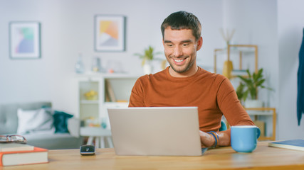 Portrait of Handsome Smiling Man Working on Laptop, Sitting at His Wooden Desk at Home. Man Browsing Through Internet, Working on Notebook from His Living Room.