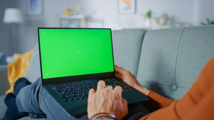 Man Lying on His Sofa At Home, Uses Laptop with Green Mock-up Screen. Over The Shoulder Shot of...