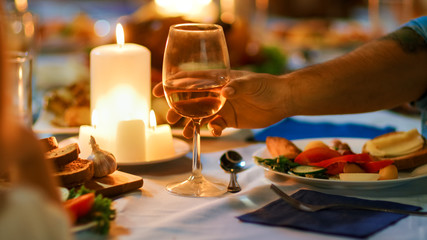 Evening Close-up Shot: Man Raises Glass at the Dinner Table and Proposes a Toast. Warm Atmosphere with Friends and Family