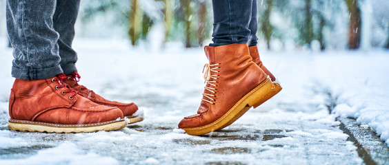 Feet of a couple on a snowy sidewalk in brown boots. Girl stands on toes while kissing. Winter...