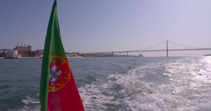 Portuguese flag waving at the wind on the back of a boat. Lisbon bridge and cityscape on the background. Shot in slow motion