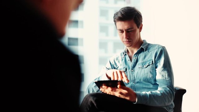Creative young business man is using tablet in the office against the background of a window and glass buildings. The camera's hiding behind a man's back. Tracking shot in slow motion.