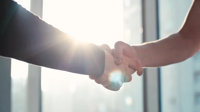Close-up of two colleagues shaking hands in the office against the background of a window and sunbeam. Tracking shot in slow motion.