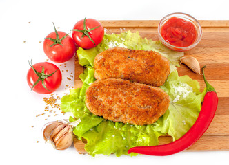Fried meatballs on lettuce on a cutting board on a white plate surrounded by tomatoes, red pepper, ketchup and garlic. View from above.