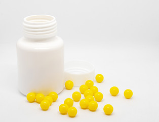 Close-up of yellow dragee vitamins scattered near a plastic open bottle on a white background.