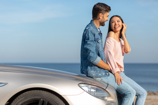 side view of caucasian boyfriend and asian girlfriend looking at each other leaning the car on sunny day