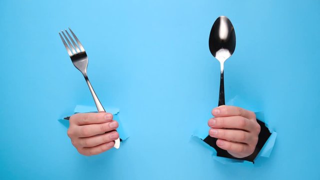Close up of female hands holding spoon and fork through torn blue paper wall. Gesture of eating dinning. Cooking, healthy homemade food concept. Copy space