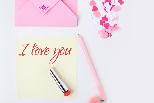 Flat lay: yellow paper, pink envelope, pen, pompom, perfume, lipstick, confetti hearts, lip print, i love you text. Making postcard in envelope for Valentine's Day.Do it yourself.Photo from the series