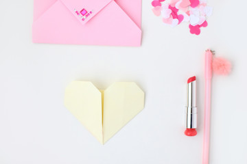 Flat lay: pink envelope, lipstick, pen, confetti hearts, yellow paper folded in the shape of heart in the origami technique. Making postcard for Valentine's Day. Do it yourself. Photo from the series