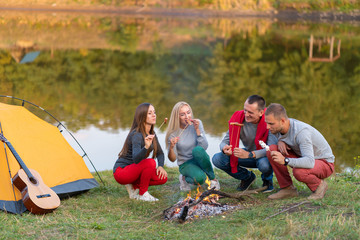 travel, tourism, hike, picnic and people concept - group of happy friends frying sausages on campfire near lake.