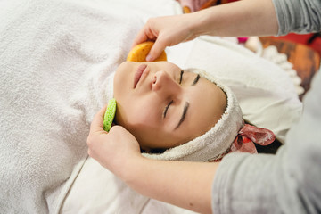 Young woman with face treatment at spa salon cosmetologist remove hold sponge mask for peeling on the face of a girl in a cosmetology room cosmetic therapy lying on bed head covered