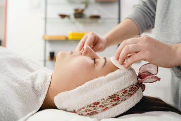 Young woman with face treatment at spa salon cosmetologist remove hold sponge mask for peeling on the face of a girl in a cosmetology room cosmetic therapy lying on bed head covered