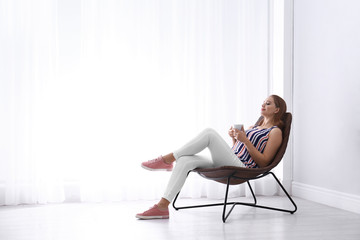 Young woman with cup of hot drink sitting in armchair at home. Space for text