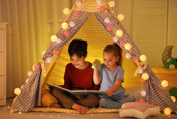 Little children with flashlight reading book in play tent at home