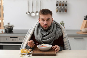 Sick young man eating tasty soup to cure flu at table in kitchen