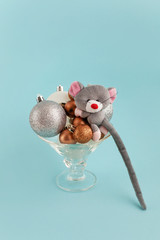 Creative colorful still life in a wide glass for cocktails on a blue background. The glass is filled with silver, gold and white shiny Christmas balls, and toy gray mouse sits on top - symbol of 2020