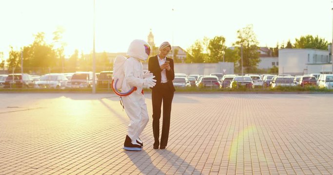 Caucasian good loking businesswoman standing at the parking and texting or tapping on the smartphone while funnt astronaut going around her and looking at the screen what she doing.