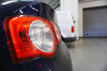 Blue luxury car in service, close-up of the rear of the car and the red taillight. In the background, the interior of the service and part of the white car are blurred.