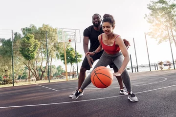 Poster Outdoors Activity. African couple girl dribbling while guy defencing backdoor on basketball court smiling happy © Viktoriia