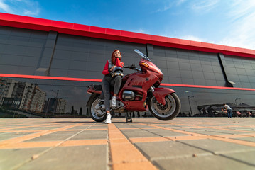 Beautiful young woman is sitting on her motobike near modern building. Red haired girl. Speed and fashion concept. Urban Lifestyle