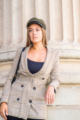 Portrait of Young East European Businesswoman in New York. Young woman with long hair, wearing patterned blazer with belt, green hat, standing against column outside office, looking forward, thinking.