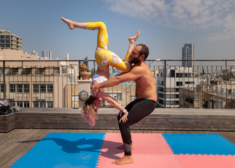 Acro yoga practice, by a muscular man and a fit woman. Shot on a rooftop with a modern urban skyline in the background.