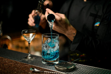 Professional male bartender pouring a blue alcoholic drink from the jigger to a measuring cup holding a glass bottle