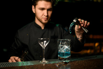 Professional male bartender pouring a blue alcoholic drink from the jigger to a measuring cup