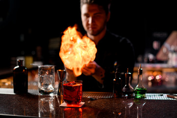 Professional male bartender serving the red alcoholic cocktail with ice cubes under the fire