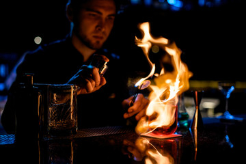 Professional male bartender serving the red alcoholic cocktail with ice fire the lemon slice on tweezers