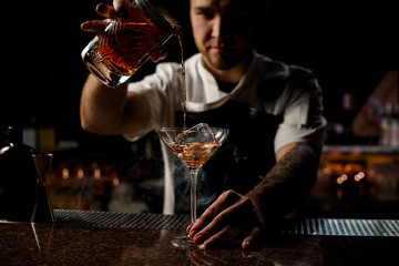 Professional bartender pouring a brown alcoholic cocktail from the measuring cup to the glass