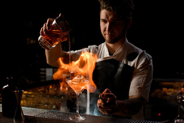 Professional bartender pouring a brown alcoholic cocktail from the measuring cup to the glass burning the fire