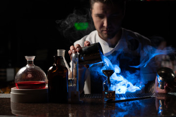 Professional male bartender pouring a transparent alcoholic drink from the steel shaker to the little bottle under blue light