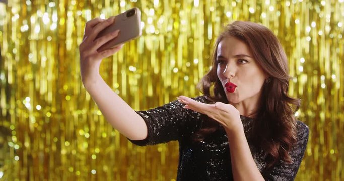 Caucasian young attractive fancy girl celebrity posing, smiling and blowing air kisses to the smartphone camera while taking selfie photos on the golden sparkling background. New Year Eve concept.