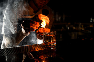 Male bartender serving the transparent alcoholic cocktail with ice fire sprinkling with the lemon...