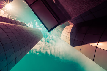 View through modern high rising skyscrapers upwards to blue sky with white clouds. Abstract architecture detail background in turquoise teal blue to burgundy purple pastel colors