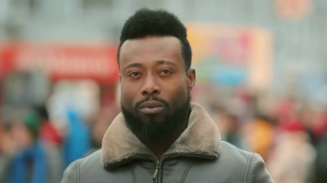 Close up African American man looking at camera standing on street city. Black afro people face portrait closeup. Confident and serious. Young adult guy. Urban background 4K. Cold winter in America.