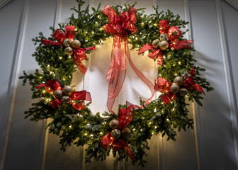 A green Christmas wreath with red ribbon bows, gold ornaments, and white lights adorn the wall of a small chapel.