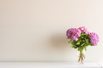 Pink hydrangea flowers with green leaves in glass vase on white side board against neutral wall...